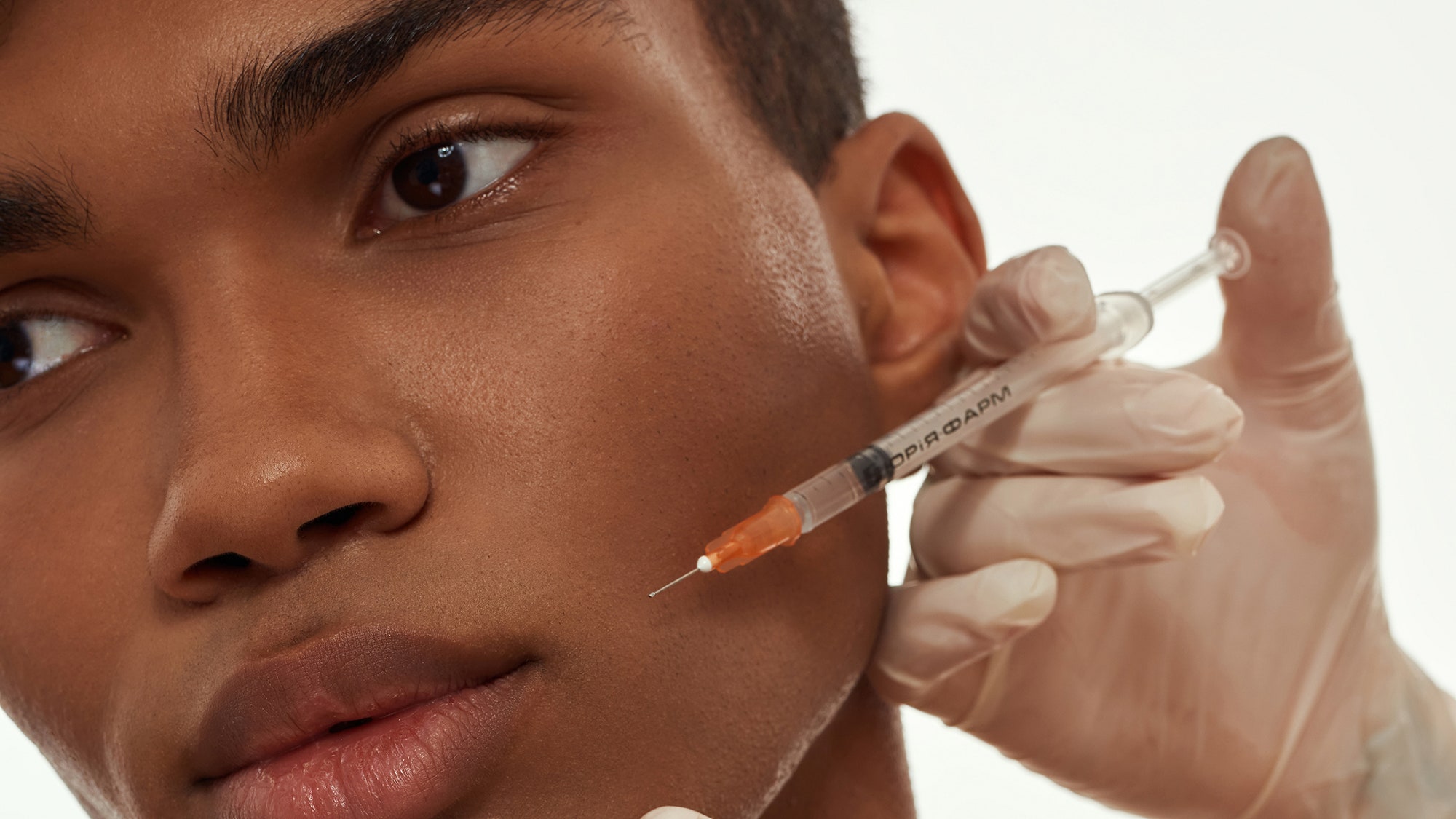 A close up of a man's face getting an injection being given by hands wearing surgical gloves against a white background