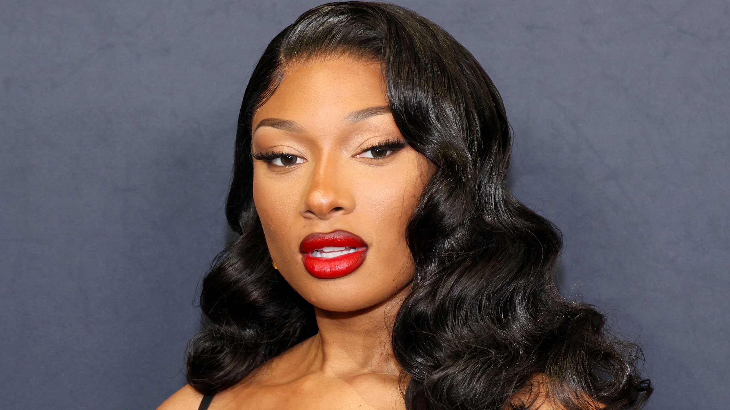 Megan Thee Stallion poses at an event in a black dress red lipstick and waves.